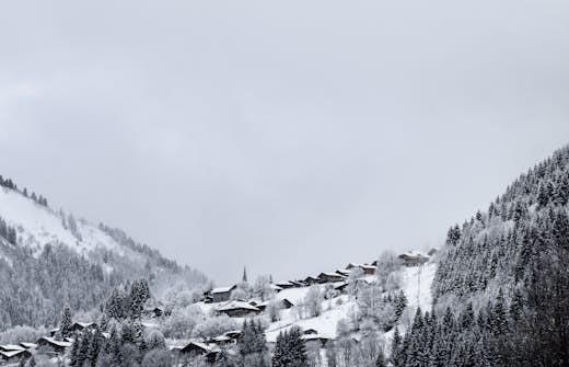 A snowy village with rustic wooden houses, surrounded by mountain peaks, viewed from behind a large rock.