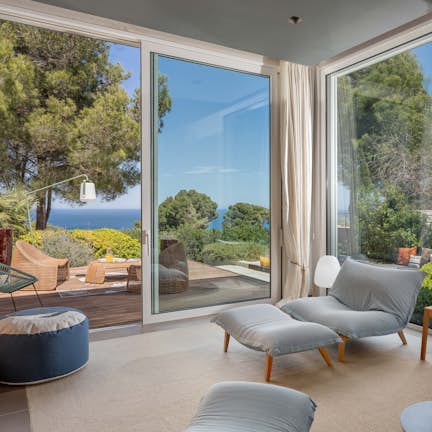 Modern living room with large glass doors opening to a terrace overlooking the sea, furnished with stylish seating and decorated with art and plants.