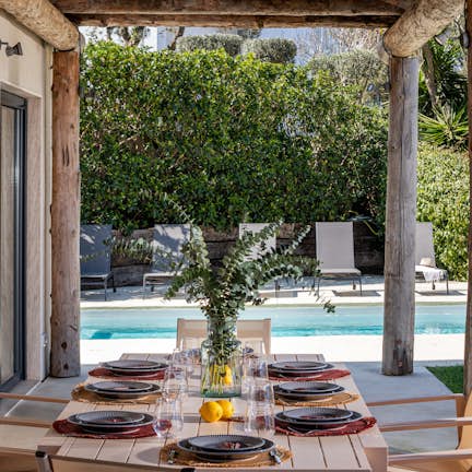 Outdoor dining area with a set table under a wooden pergola, overlooking a swimming pool surrounded by lush greenery.