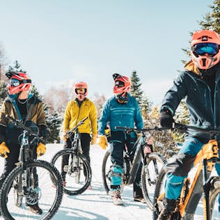 2022_les-2-alpes-mi-avril-pyrene-duffau-12 Four cyclists in colorful gear and helmets ride mountain bikes on a snowy path, surrounded by evergreen trees and a clear sky.