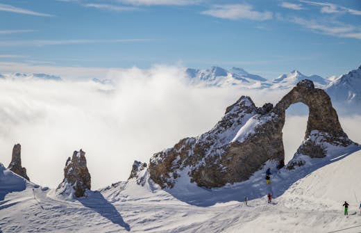 Alpine landscape featuring rocky arch with skiers on snow-covered slopes above cloud level, under a clear blue sky.