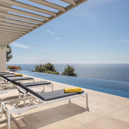Luxury house with an infinity pool overlooking the ocean, featuring clear skies and sun loungers on a tiled patio.