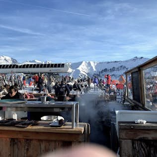 Outdoor mountain restaurant busy with diners, featuring a food counter in the foreground and snowy peaks in the background under a clear blue sky.