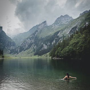 A person swimming in a clear mountain lake with towering cliffs and lush greenery under a cloudy sky.
