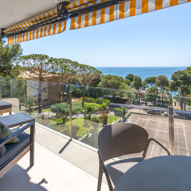 Costa Brava accommodation - Apartment Areca - Outdoor patio with wicker furniture, dark cushions, and green pillows. Glass railing, a coffee table, and a window are also visible. Bright, sunny weather.