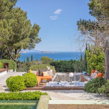 Villa Verde in Begur, Costa Brava Luxurious modern house with a large pool, surrounded by lounge chairs and umbrellas. The two-story building features large glass doors, beige walls, and a lush garden.