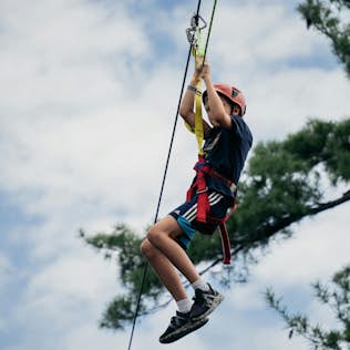 Tree-top Tree Climbing Course A child wearing a helmet and harness rides a zip line outdoors, with trees and a cloudy sky in the background.