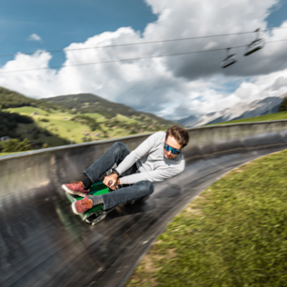 A person in sunglasses and a casual outfit sliding down a steep, curved alpine slide against a scenic mountain backdrop.
