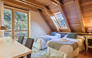 Vielha - 4 Baqueira Beret alojamiento - Casa Montcorbison - A cozy attic bedroom with two single beds, a skylight, wooden walls, a desk with chairs, and a window showing trees and mountains outside.