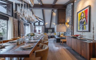 Morzine alojamiento - Chalet Coquelicot - Chalet Coquelicot, Courchevel Morzine alojamiento - Chalet Coquelicot - Elegant dining room with a long wooden table, modern chandelier, stone fireplace, and artwork on the wall. Large windows with curtains.