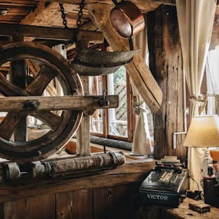 Rustic wooden interior featuring a vintage typewriter, a large wooden wheel, and decorative items on a timber ledge by a window with sheer curtains.