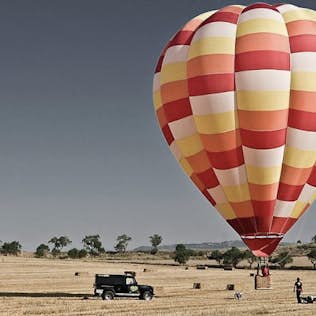 A colorful hot air balloon hovers above a golden field with hay bales; a black suv and people are visible below.