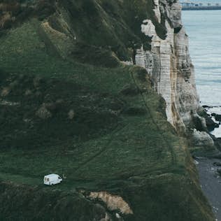 A camper van parked on a grassy cliff overlooking the sea at dusk, with steep rocky cliffs descending to the water.