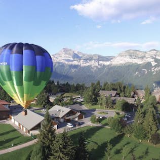 A hot air balloon floats above a small village surrounded by mountains and forests under a clear sky.