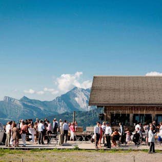 A group of people gathering outside a mountain hut with a cable car nearby, surrounded by alpine scenery under a clear blue sky.