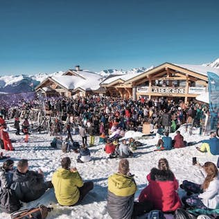 Crowd gathered at a ski resort enjoying an event with snow-covered mountains in the background, under a clear blue sky.