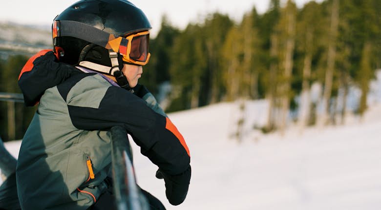 A person wearing a helmet and ski goggles leans on a rail, overlooking a snowy landscape with trees in the background.