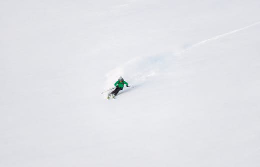 A skier in a green jacket descending a snowy slope with fresh powder trailing behind.