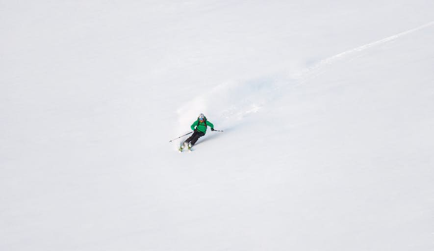 A skier in a green jacket descending a snowy slope with fresh powder trailing behind.