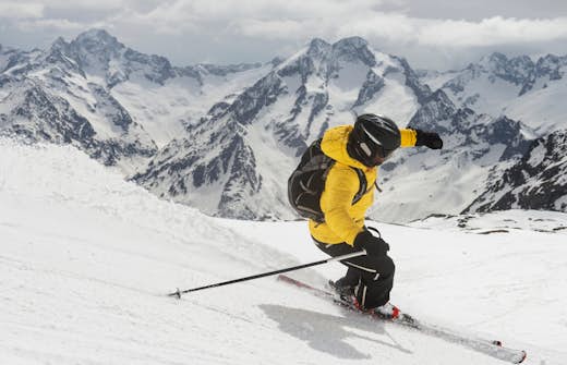 A skier in a yellow jacket carving a turn on a snowy mountain slope with rugged peaks in the background