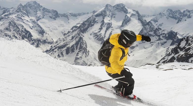A skier in a yellow jacket carving a turn on a snowy mountain slope with rugged peaks in the background.