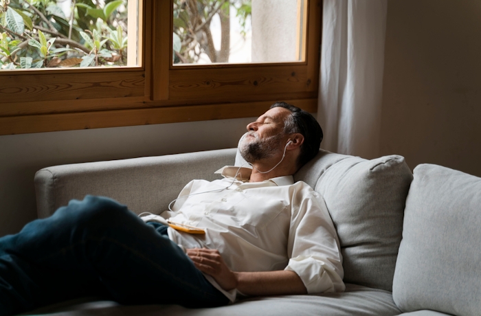 Man relaxing on a couch with headphones, eyes closed, near a window with a view of greenery.