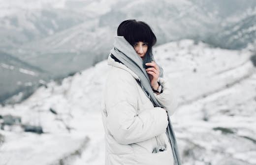 A woman in a white coat and scarf stands in a snowy landscape, looking towards the camera.