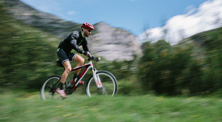 A cyclist in a black outfit and red helmet rides a mountain bike on a grassy trail with blurred green scenery in the background, suggesting motion.