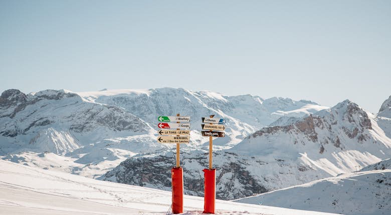 Snow-covered mountain landscape with ski direction signs pointing various routes and destinations. The sky is clear, and distant snow-capped peaks are visible.