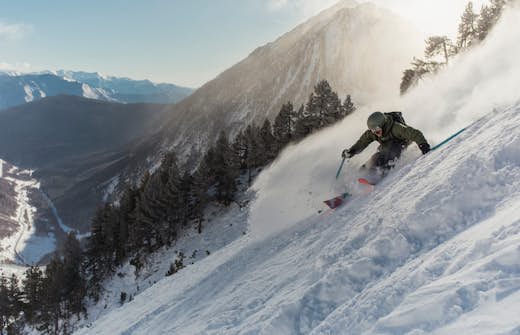A skier aggressively carving through powder snow on a steep mountain slope with scenic mountain views in the background.