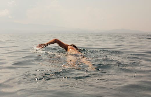 A person swimming freestyle in a calm sea with mountains visible in the distant haze.