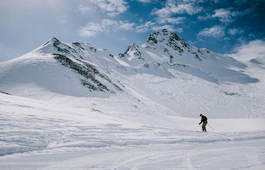 A skier descends a snowy slope with towering, sunlit mountains in the background under a clear blue sky.