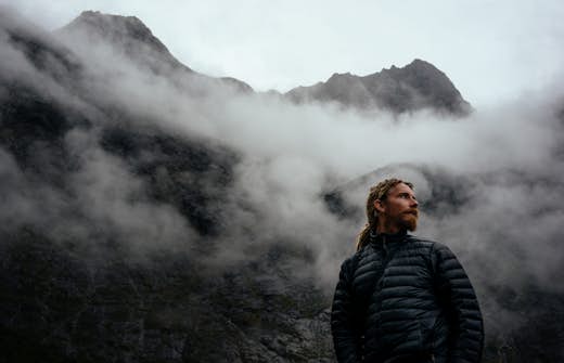Man with a beard wearing a puffy jacket looks contemplatively to the side, with misty, rugged mountains in the background.
