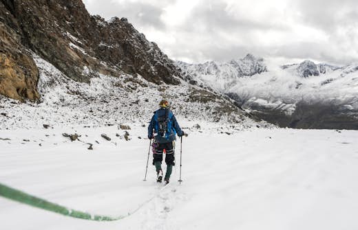 A hiker wearing a blue jacket and carrying a backpack treks through snow towards distant mountains under a cloudy sky.