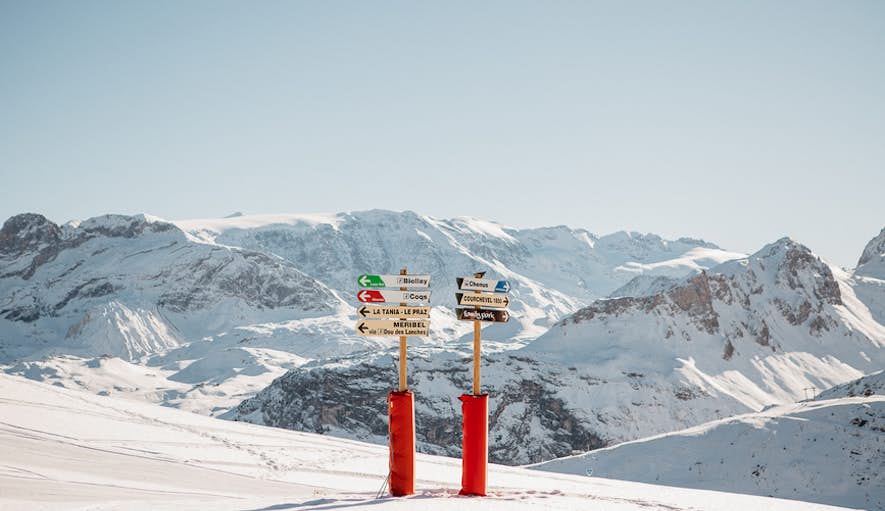 Directional signs on a ski slope with snow-covered mountains in the background under a clear blue sky.