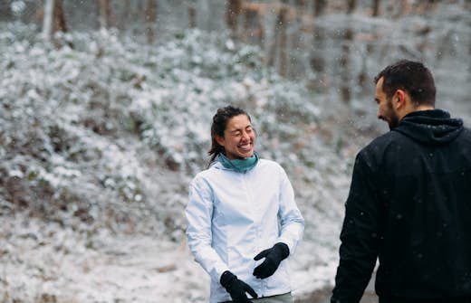 A woman in a white jacket laughs with a man in a black jacket in a snowy forest setting.