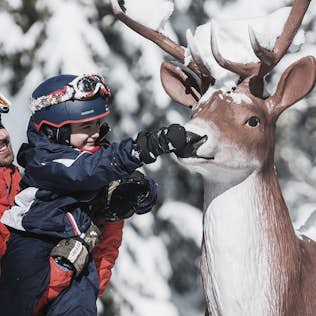 A man holding a child who is playfully touching the nose of a life-sized model deer in a snowy setting. both are dressed in winter gear.