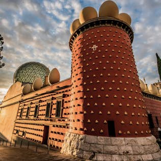 An image of the unique salvador dali theatre-museum in figueres, spain, showcasing its distinctive red facade with rounded shapes and large eggs on the roof at sunset.