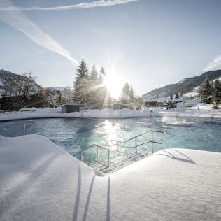 Outdoor swimming pool surrounded by snowy landscape with the sun shining brightly in a clear blue sky.