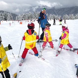 A ski instructor stands with a group of children in ski gear and bright yellow bibs on a snow-covered slope.