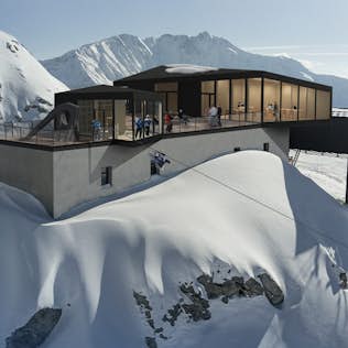 Modern mountain lodge with large windows and a balcony, surrounded by snow-covered slopes, with people enjoying the view.