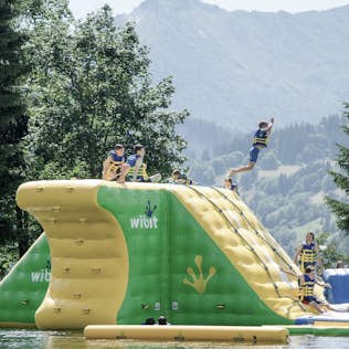A child jumps from a large inflatable water slide into a lake, with others watching, set against a mountainous backdrop.
